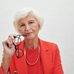 Menu A senior woman with white hair and eyeglasses in a red blazer holding spectacles thoughtfully.